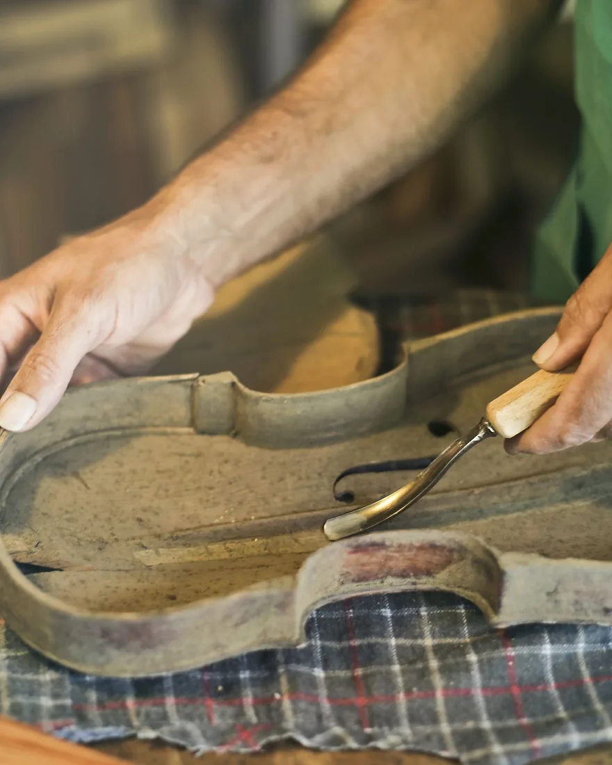 Manos trabajando con una gubia pequeña sobre el fondo de un violín antiguo apoyado en un paño